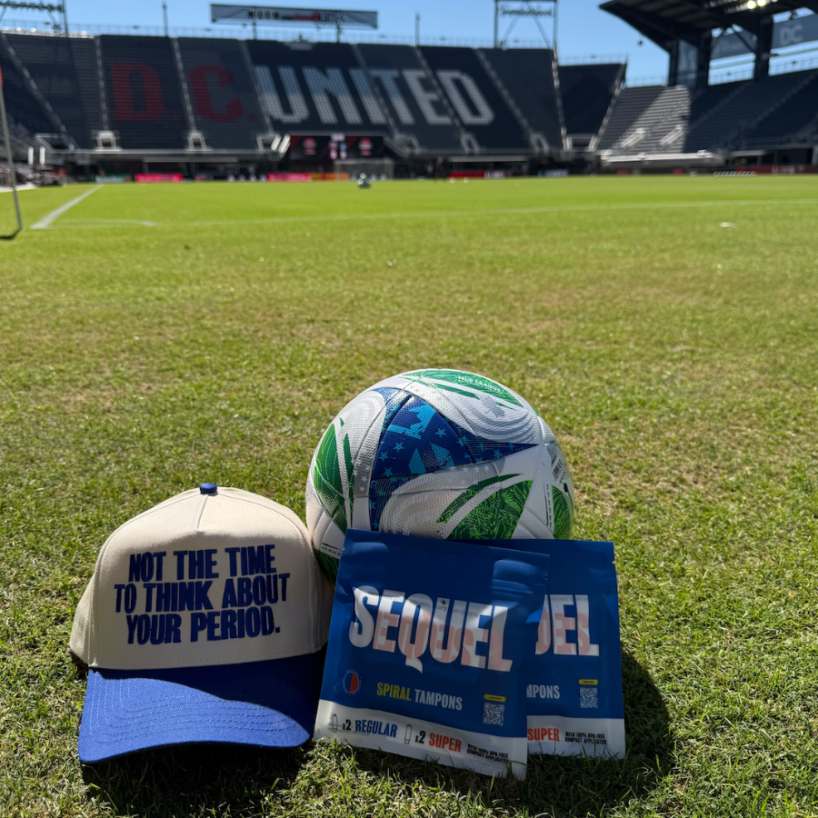 A blue and beige baseball cap with the text 'NOT THE TIME TO THINK ABOUT YOUR PERIOD.' sits on a green soccer field next to a soccer ball and a blue 'SEQUEL 27' sign, with a stadium in the background.