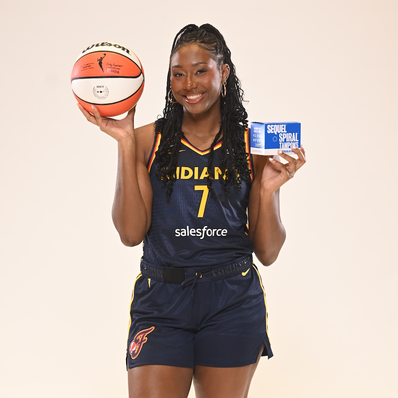 A woman stands against a plain light beige studio background wearing a navy basketball uniform with yellow lettering and shorts, holding an orange basketball in one hand and a small blue box in the other. Visible text on the uniform includes 'INDIANA', '7', and 'salesforce'; the basketball also shows Spalding branding.