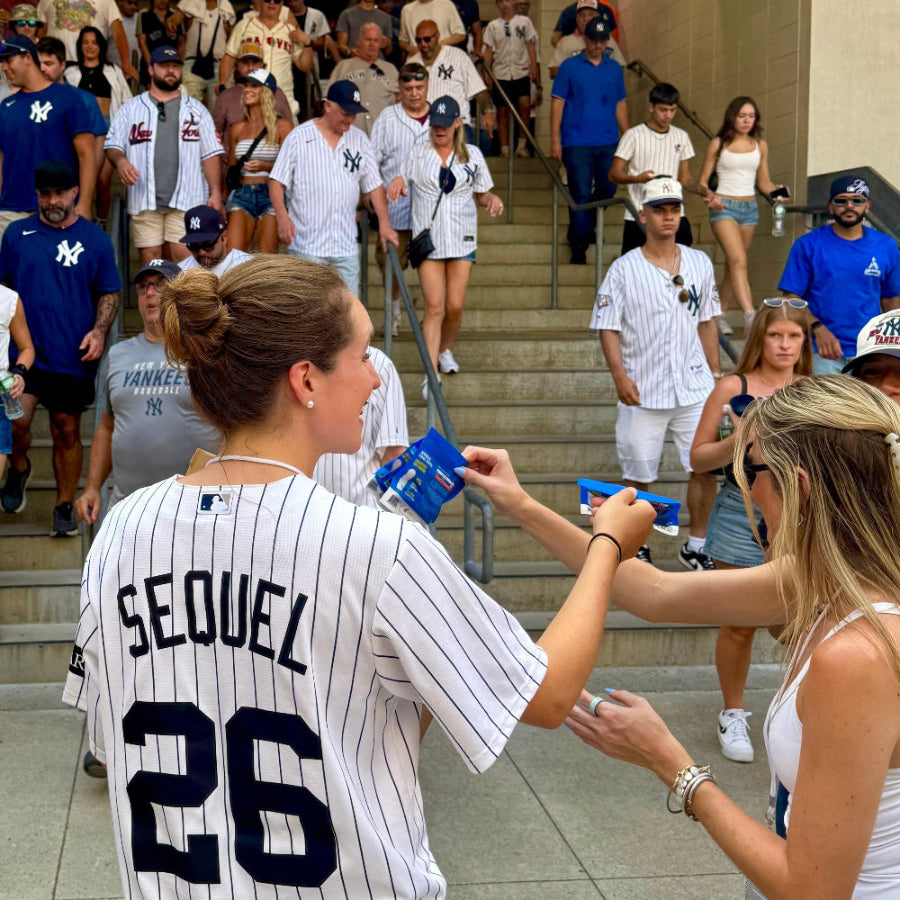 A woman in a white pinstripe New York Yankees jersey with 'SEQUEL 26' on the back stands on outdoor stairs, signing autographs with a blue microphone while a crowd of fans in matching team apparel walks up the steps behind her.