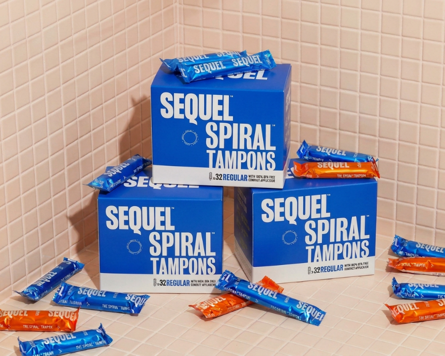 Three blue boxes of Sequel Spiral Tampons arranged on a tiled surface with several individually wrapped tampons scattered around. The packaging is predominantly blue and white, with some orange-colored tampons visible in the wrappers.