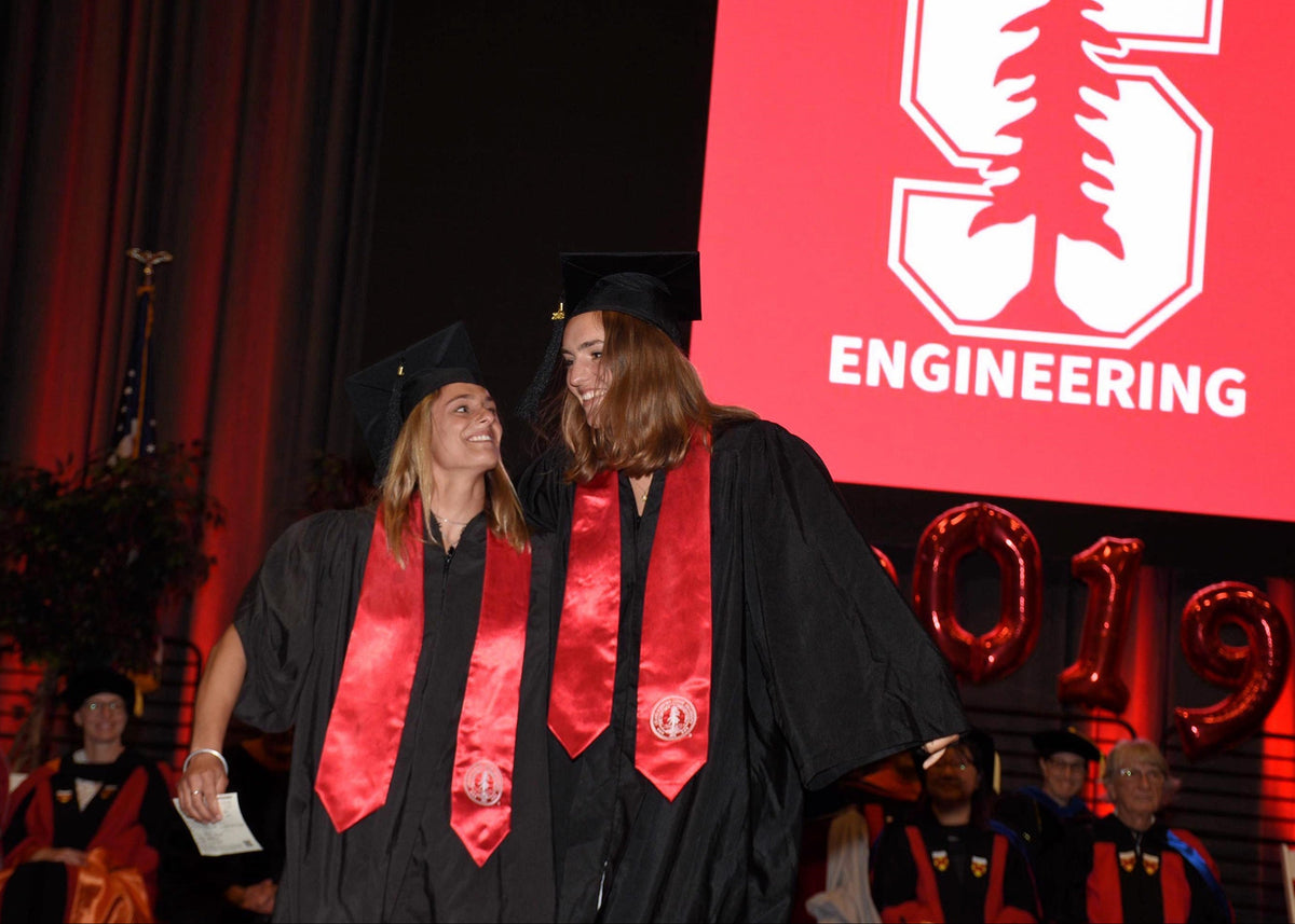 Two graduates in black caps and gowns with red satin stoles walk on stage in front of a large red screen that reads 'S ENGINEERING' and '2019'.